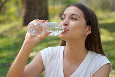 Şişeden su içen sağlıklı genç bir kadın. Brezilya 'lı beyaz kadın şehir parkında çimlerin üzerinde oturmuş su içiyor..