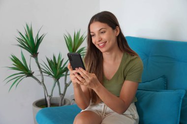 Evde akıllı telefonla internetten alışveriş yapmak. Çevrimiçi sipariş veren mutlu genç kadın.