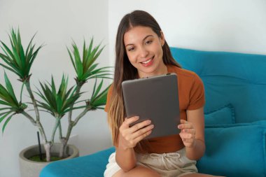Tablet kullanan İspanyol genç bir kadının portresi dairesinde kanepede oturuyor.