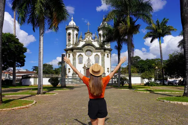 Brezilya Koloni Mimarisi. Brezilya 'nın Minas Gerais kentindeki Sao Joao del-Rei kasabasındaki Assisi kilisesinin önünde kollarını kaldıran turist kız..