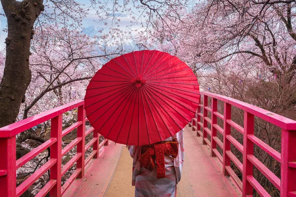 Woman traveller with a red umbrella and walking over the bridge with Fuji mountain and Sakura flower background in Tokyo city, Japan