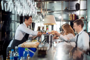 Asyalı barmen ve müşterisi barın tepesinde ve restoranda içki tezgahıyla konuşuyor.