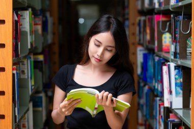 Asyalı kadın öğrenci, üniversite kütüphanesinde ders kitabı okuyor ve ev ödevi yapıyor.