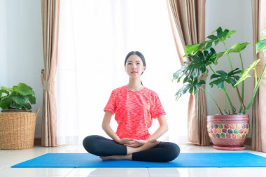 Asyalı kadın evinde yoga minderiyle oturma odasında egzersiz yapıyor.