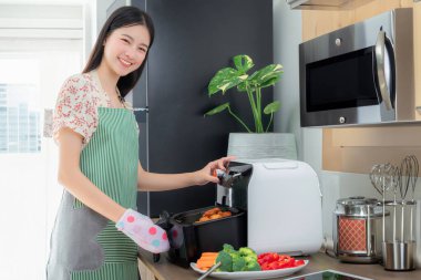 Asyalı eş gülümseyerek ve kızarmış tavuk pişirerek evdeki mutfağında elektrikli fritözünü kullanıyor. 