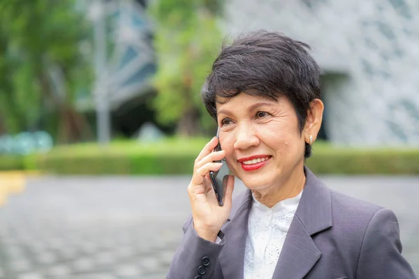 Asian old businesswoman call the mobile phone with her customer infront of her company office with smile and expression