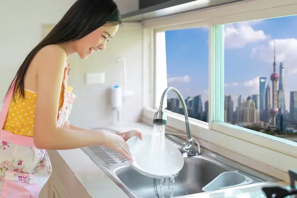 亚洲女人在她的公寓里做洗碗工作 窗户上挂着中国城市的景观 图库照片