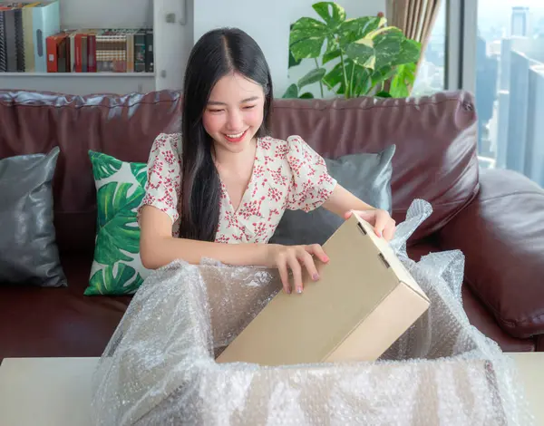 アジアの女性は幸せで 彼女のマンションの街の背景を持つ彼女のリビングルームのソファーのオンラインショッピングからパッケージを解放 ロイヤリティフリーのストック写真