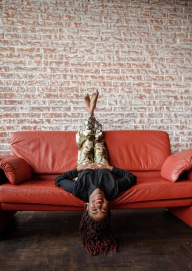 Neşeli genç siyahi kadın kırmızı deri koltukta baş aşağı uzanmış kameraya bakıyor ve duvarla odada gülümsüyor. Mutluluk ve neşe kavramı. Çocuk gibi davranarak eğleniyorum.