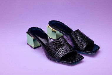 Eflatun bir arka planda izole edilmiş metalik topuklu, göz alıcı siyah sürüngen deri parmak arası terlikler. Şık parti ayakkabıları.