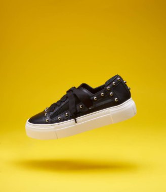 Tekstil işlemeli modern siyah deri ayakkabı, gümüş perçinler ve parlak sarı arka planda fotokopi alanı ile uçan tıknaz beyaz dikişli taban. Havalandırma konsepti, yaşam tarzı ürün fotoğrafı.