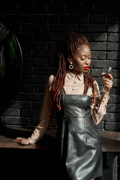 Attractive African girl with bold makeup and dreads standing against a black wall in the loft interior and holding a glass of white wine while looking down. Concept of glamorous life