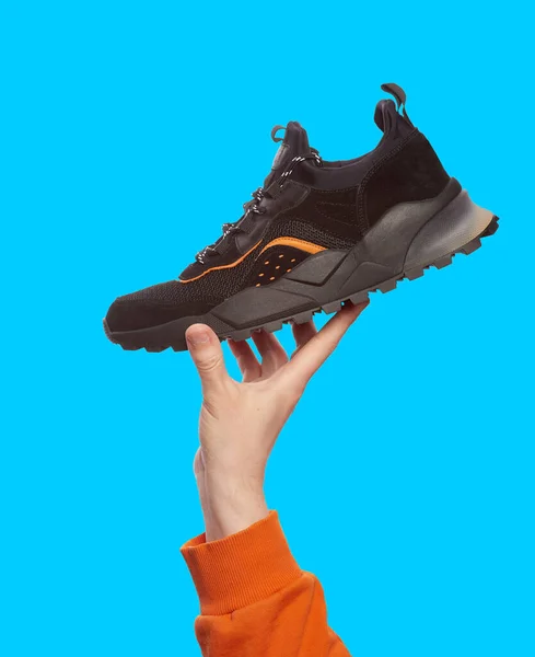 Stylish black sneaker with chunky sole in man\'s hand, isolated on a bright blue background. Mock-up for advertising a sports footwear shop