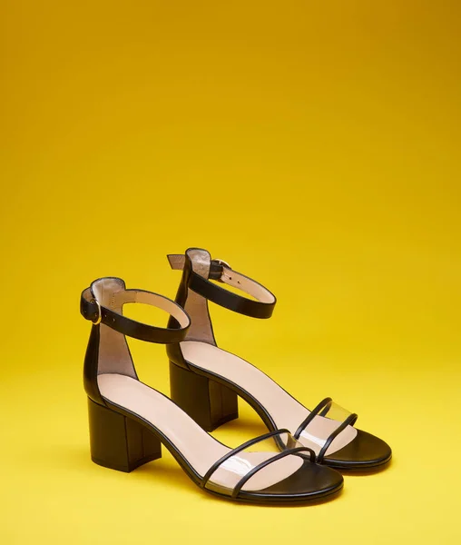 Fashionable open-toe black women's shoes with block heels, clear vamps and ankle straps isolated on a bright yellow background with copy space on top. Side view. Concept for a modern shoe store.