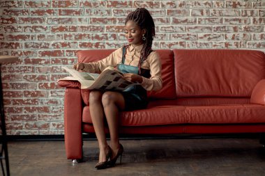 Çekici bir Afrikalı kadın kırmızı koltukta oturuyor ve gazetede iyi haberler okuyor. Fotokopi alanı olan tuğla bir arka plan duvarına karşı. İçerik yazarı, yardımcı editör, ilişki yöneticisi, muhabir