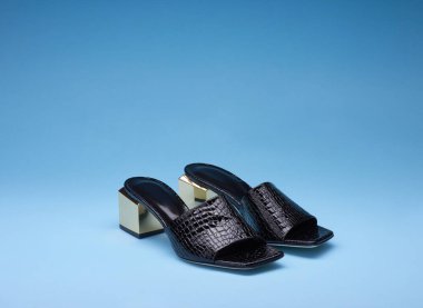 Süslü kadınların açık parmaklı siyah terlikleri. Timsah derisi dokular ve altın metalik topuklar. Gradyan mavi arka planda izole edilmiş. Modern ve moda bir ayakkabı mağazası..