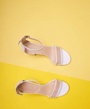 Süslü topuklu beyaz kadın sandaletleri, temiz vampirler, ayak bileği kayışları ve sarı arka planda izole edilmiş bej tabanlar. Üst manzara, düz yatış. Moda fotoğrafçılığı, yaz sezonu ayakkabı konsepti.