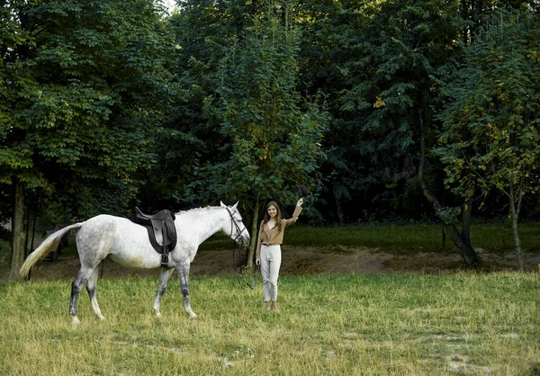 An attractive girl standing with a saddled grey horse on a grass field with trees area behind and taking off her hat, making a greeting. Spending a weekend in nature with a pet horse. Horse therapy