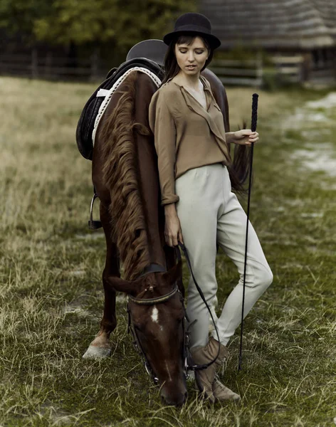 一个戴着黑色帽子的年轻妇女正在放牧一匹棕色的马 一个女人和她的马在乡间的草地上 — 图库照片