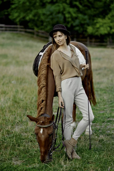 头戴黑帽的女人站在草地上放牧着棕色的马 女人和她的马拴在草地上 — 图库照片