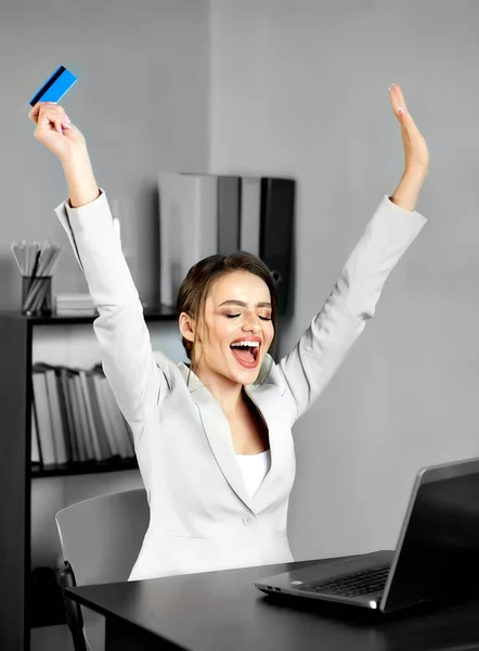 Attractive cheerful woman with her hands raised and eyes closed, holding a bank credit card while sitting at the table with a laptop. Excited, happy, winner. Celebrating successful shopping deal.