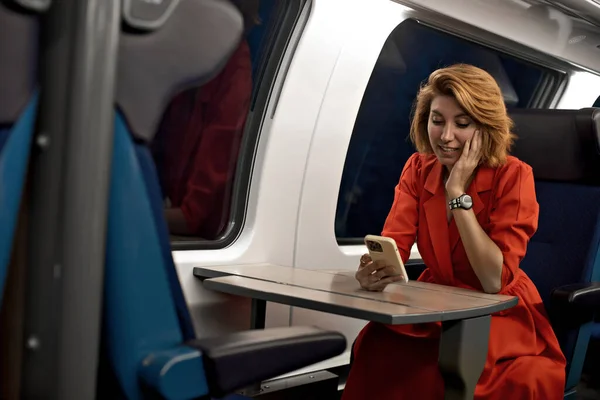 Smiling woman using mobile phone device by window with reflection on glass in train window at her wagon. Online mobile app with free wi-fi in train for transport leisure entertainment.