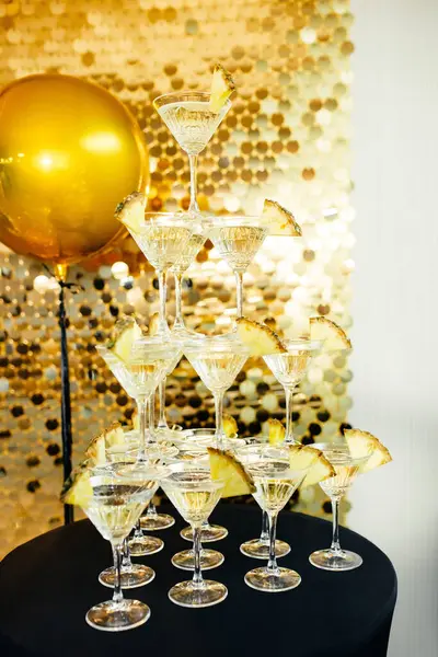 Tower made of glasses with champagne on table indoors. Decorated with pieces of pineapple in form of a pyramid with four rows on golden background of shiny sequins and large golden inflatable ball.