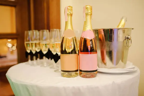 白とピンクのシャンパン2本が丸いテーブルの上に立っていて その背後にはドリンクとアイスバケツがついているメガネがあります 職場での企業のお祝い 結婚式 誕生日 クリスマスパーティー ロイヤリティフリーのストック写真