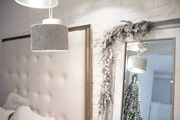明るいモダンなベッドルームの鏡は 雪に覆われた枝のガーランドで ツインリングライト付きのクリスマスツリーを反映しています ベッドの柔らかいヘッドボードと現代的なランプ ロイヤリティフリーのストック写真