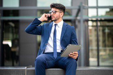 Genç yönetici tabletini kullanarak modern bir şehirde bir bankta oturuyor ve telefonla konuşuyor.