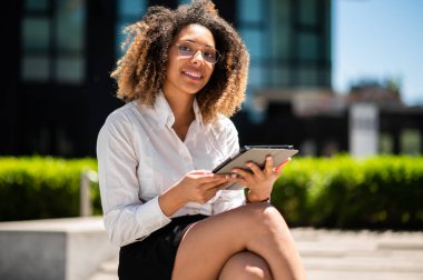 Gülümseyen Afro-Amerikan iş kadını dijital tablet kullanarak bankta oturuyor.