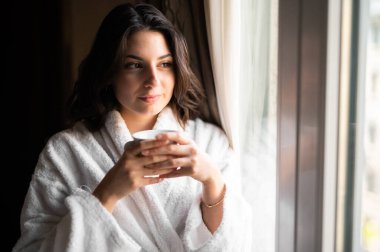 Genç bir kadın bornoz giyiyor ve bir otel odasının penceresinin yanında kahve içiyor.
