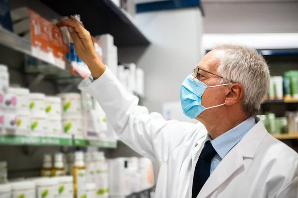 Senior pharmacist searching for a product on a shelf in his store, wearing a mask due to coronavirus