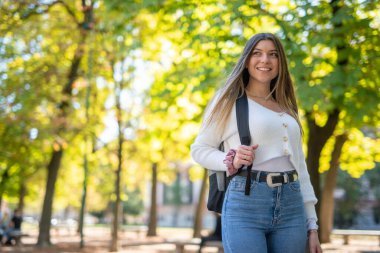 Üniversitenin yakınındaki parkta yürüyen gülümseyen bir üniversite öğrencisi.