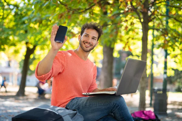 Hombre Joven Usando Una Computadora Portátil Parque Mientras Muestra Teléfono —  Fotos de Stock
