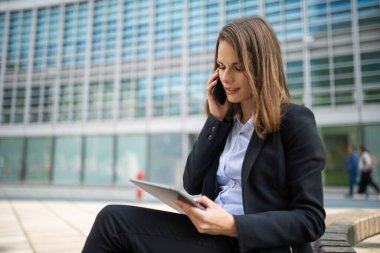 Elinde tabletle cep telefonunu kullanan gülümseyen genç bir iş kadını.