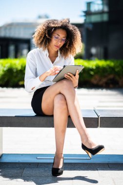 Gülümseyen Afro-Amerikan iş kadını dijital tablet kullanarak bankta oturuyor.