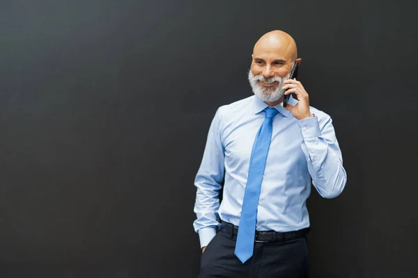 電話で話している間に黒い壁の横に立っているひげそりのビジネスマン — ストック写真