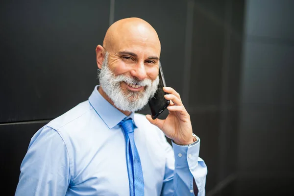 電話で話している間に黒い壁の横に立っているひげそりのビジネスマン — ストック写真