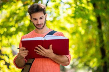 Genç bir erkek öğrenci açık havada kitap okuyor.