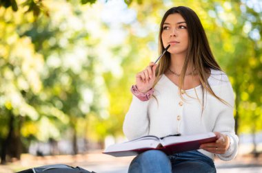 Güzel bir üniversite öğrencisi parkta bir bankta kitap okuyor.