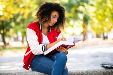 Güzel Afrikalı üniversite öğrencisi parkta bir bankta kitap okuyor.