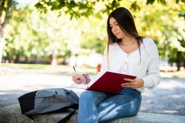 Güzel bir üniversite öğrencisi parkta bir bankta kitap okuyor.