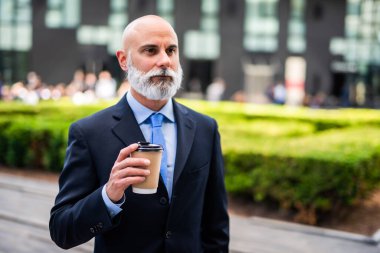 Beyaz sakallı kel bir işadamı şehirde yürürken kahve içerek konuşuyor.