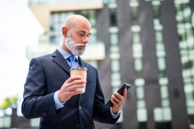 Beyaz sakallı kel bir işadamı telefonda konuşuyor ve kahve içiyor.