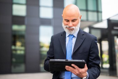 Modern bir şehirde beyaz sakallı kel bir işadamı tabletini kullanıyor.