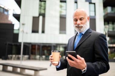 Üst düzey kel yönetici, beyaz sakallı, dışarıda kahve içerken akıllı telefonunu kullanıyor.