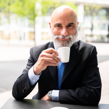 Beyaz sakallı, son sınıf kel menajer dışarıda kahve içiyor.