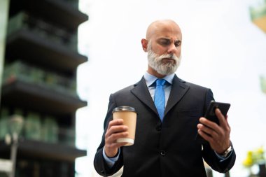 Üst düzey kel yönetici, beyaz sakallı, dışarıda kahve içerken akıllı telefonunu kullanıyor.