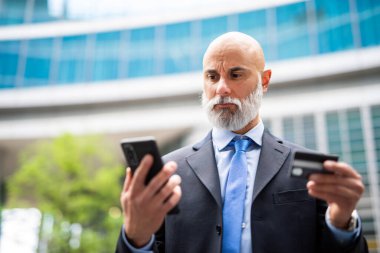Yakışıklı, şık, kel bir müdür. Açık havada akıllı bir telefonla kredi kartıyla bir şeyler alıyor.
